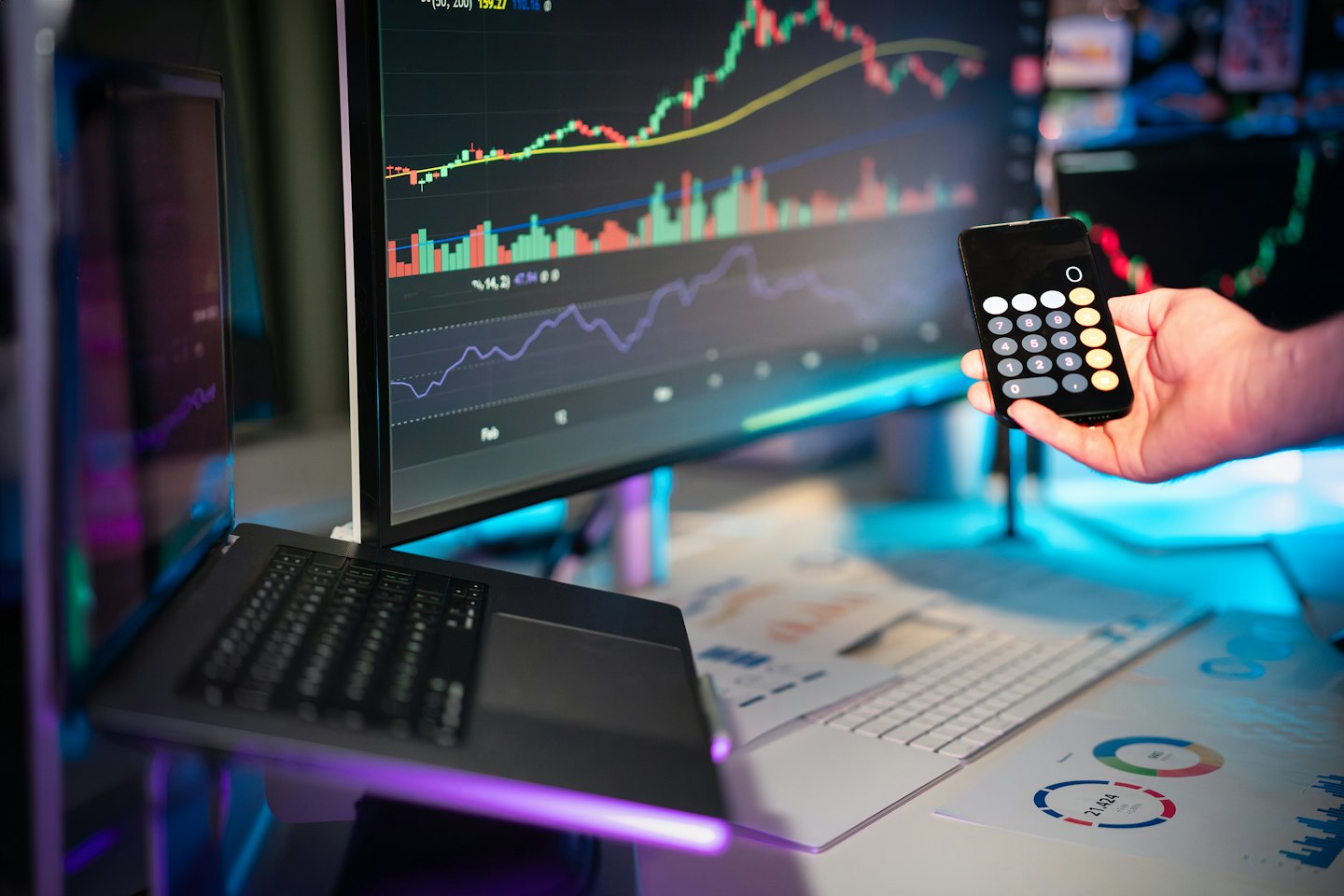 Hand holds phone showing calculator near financial data on monitors.