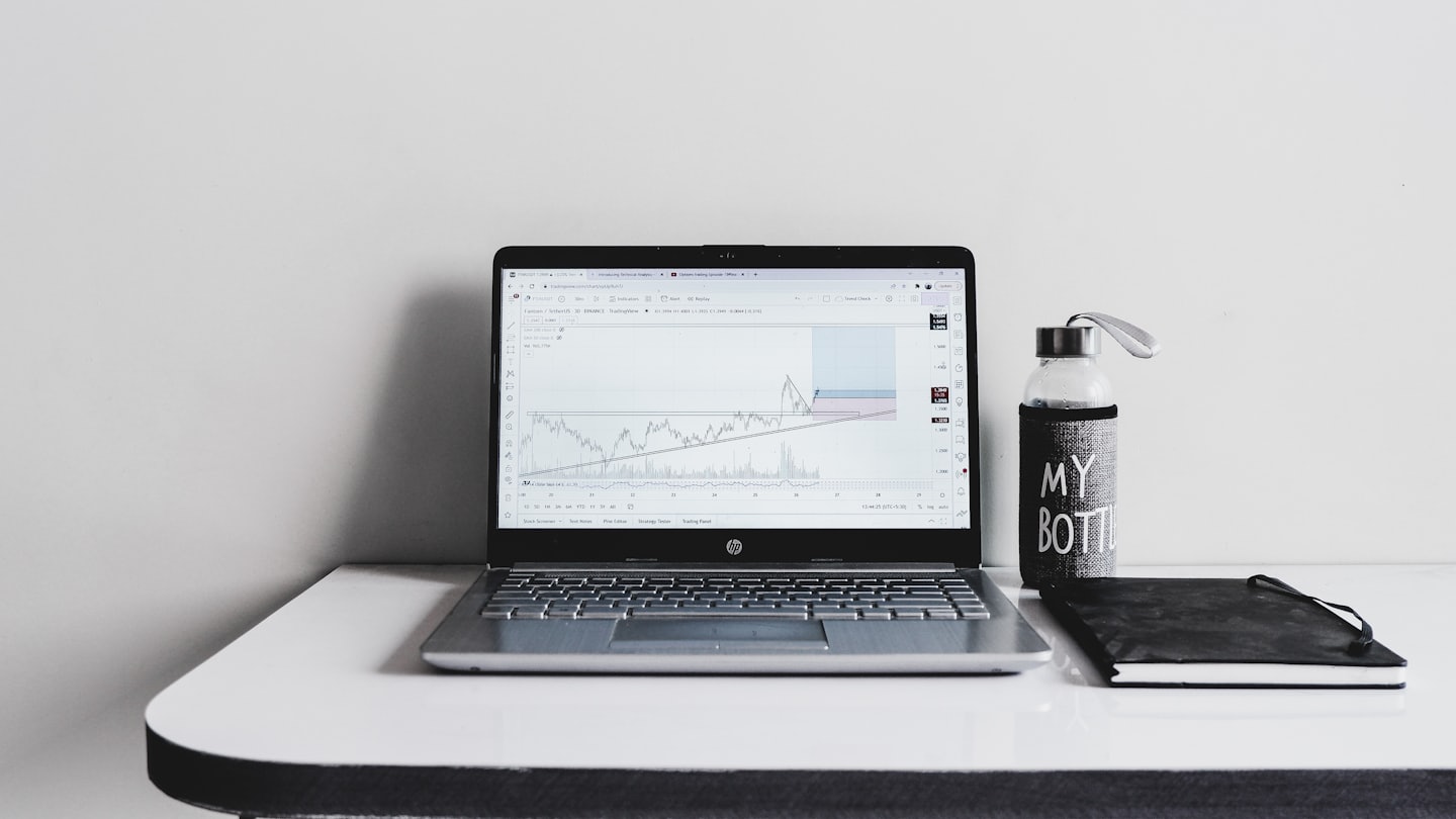Laptop with financial charts sits beside a notebook and water bottle.